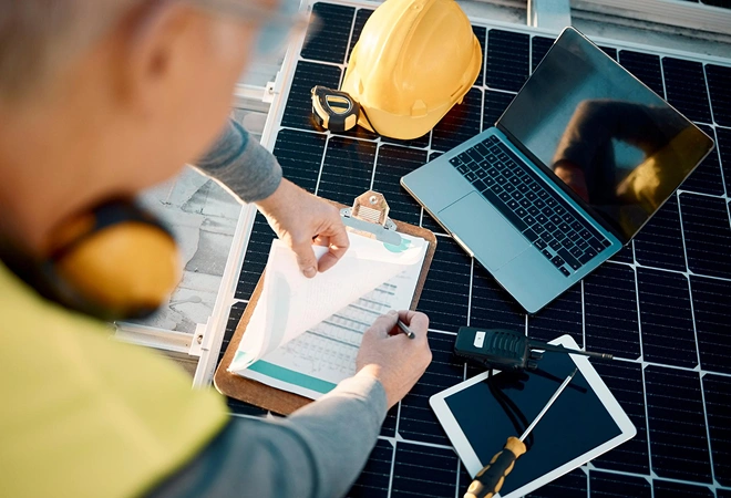 Person mit Klemmbrett, Laptop und Tablet arbeitet an einem Solarpanel